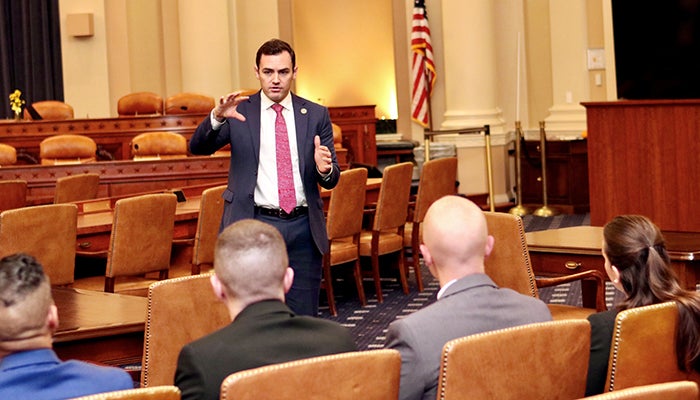 A man in a dark suit and pinkish necktie addresses a row of people in brown chairs.