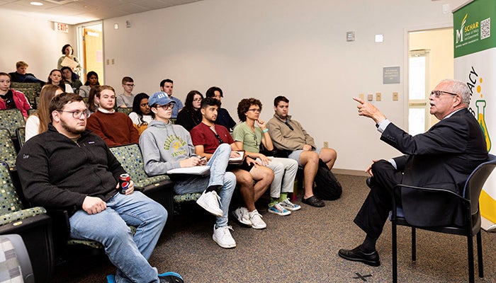 A man in a dark suit with gray hair and a moustache sits in front of a Democracy Lab and points to students in front of him.
