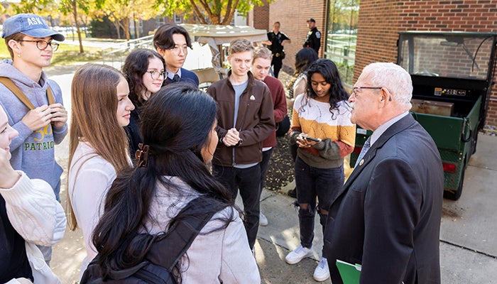 A man in gray hair and moustache is surrounded by college students out doors.
