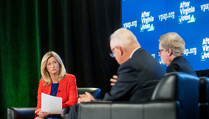 A blonde woman in a red blazer over a blue blouse holds a sheet of paper and looks at two men in chairs across from her.