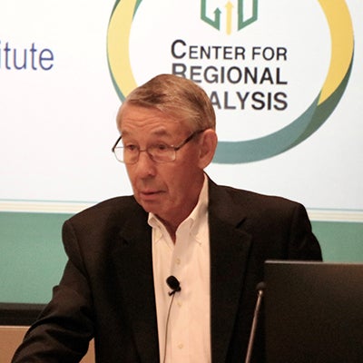 A man in a suit stands at a podium with a slide on a screen behind him.