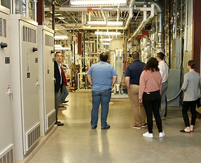 A group of people with their backs to the camera gaze at a room of pipes and lights.