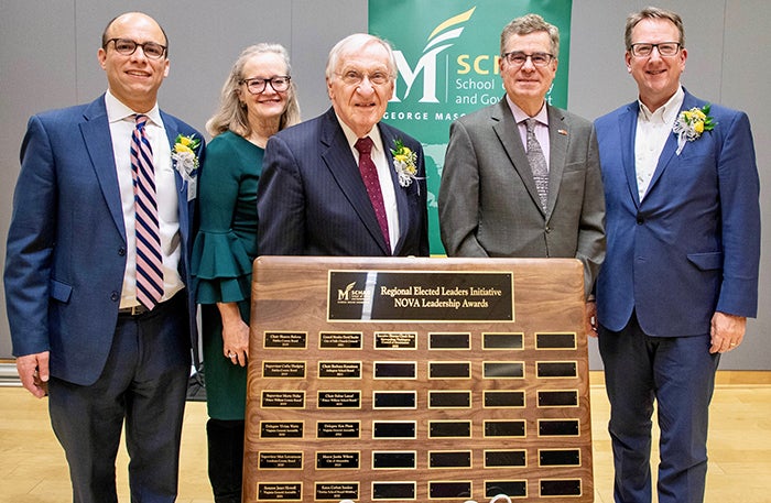 Five people dressed for success stand in front of a wooden plaque.
