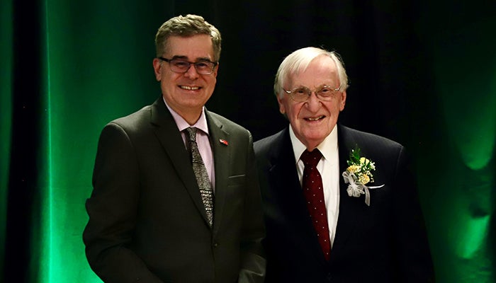 Two men in suits, one with a corsage, pose in front of a green curtain.