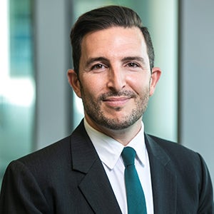 A man in a dark suit and tie with a short beard smiles at the camera.