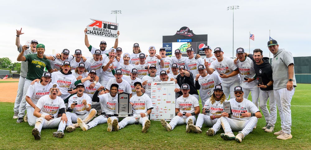 Mason's Baseball team is positioned, some sitting and some standing, on a baseball field, cheering. One player holds a sign that reads "A10 Baseball Champions." Another holds their trophy while a third holds a poster of the bracket of games they played through to win.