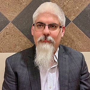 A man with a long white goatee beard and moustache and wearing an earring and glasses gazes to his right.