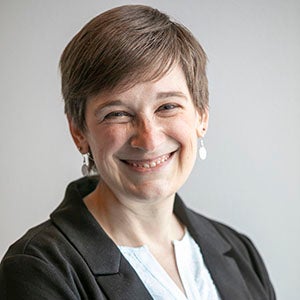 A woman in a dark jacket and light blouse with short brown hair smiles at the camera.