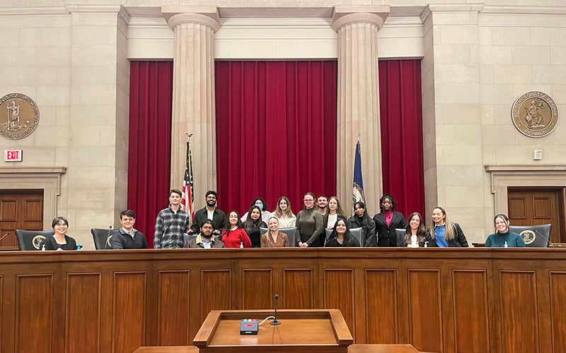 A group of students sit and stand behind a wooden dais and in front of a red curtain and two large pillars.
