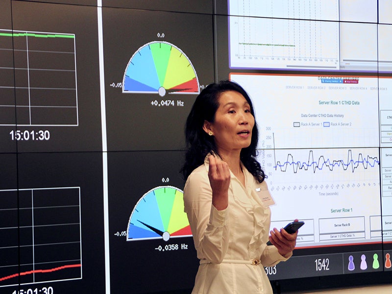 A woman with dark hair in a cream-colored dress stands in front of several panels of screens showing charts, grids, and data.