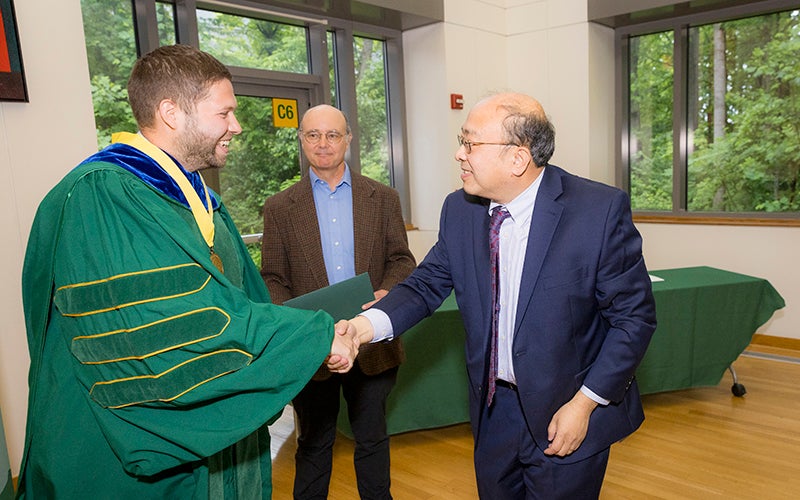 A man in a green robe with a yellow ribbon around his neck shakes hands with a man in a blue suit while a man in a brown jacket watches.