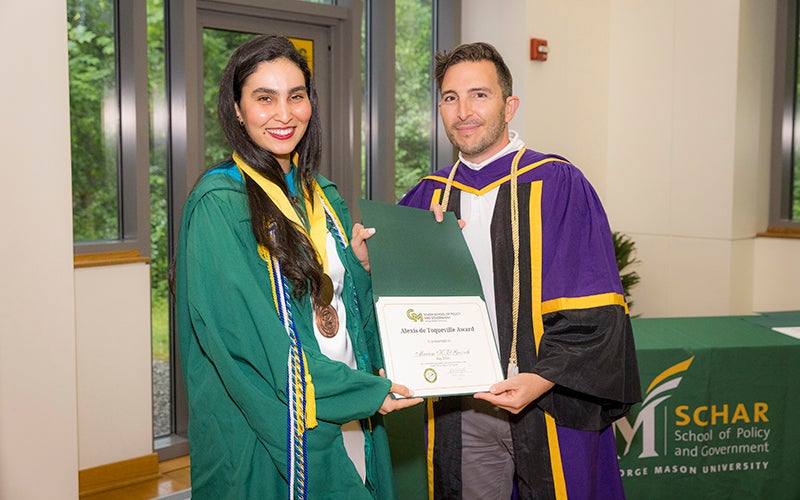 A woman in a green robe with a yellow ribbon around her neck holds a certificate with a man in a purple robe.