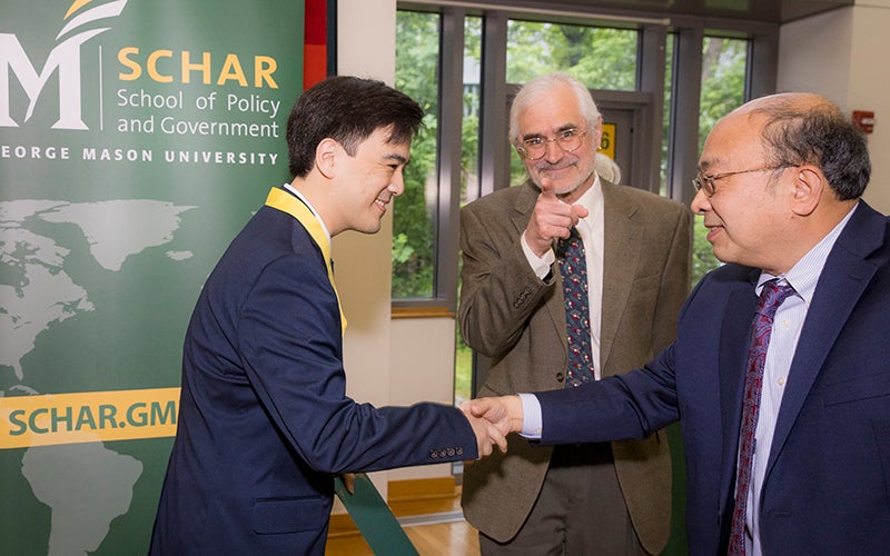 A man in a blue jacket wearing a yellow ribbon shakes hands with a man in a blue jacket while a man in a brown jacket smiles and points to the camera.