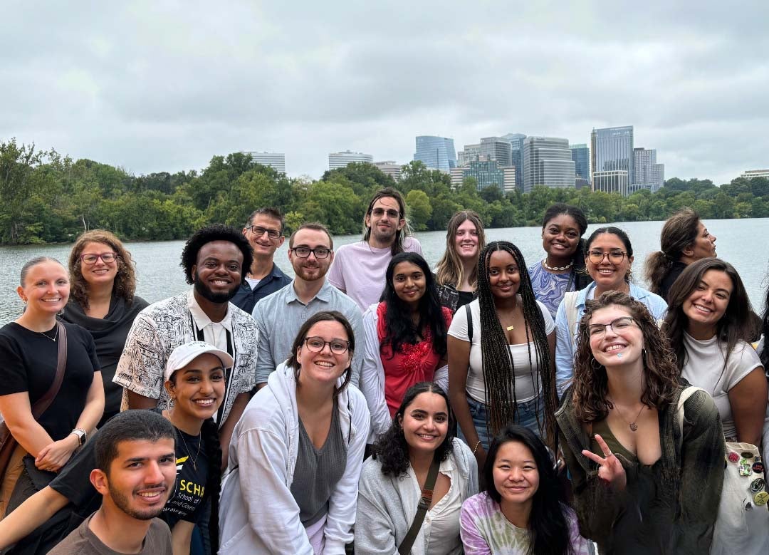 A large group of men and women pose on a boat with the Potomac River and office buildings in the background.