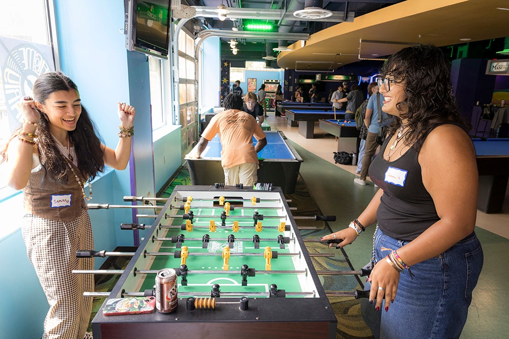 Two young women, one with her arms raised, stand opposite a foosball table and smile.