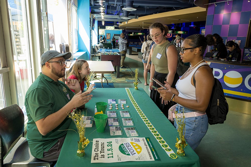A young man wearing a baseball cap and a young woman sit at a table and talk to two women who are standing on the other side.
