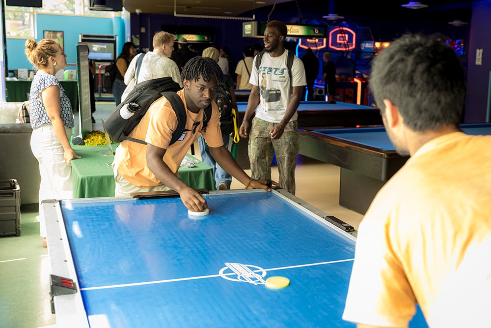 Two young men, one wearing a backpack, play air hockey.