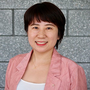 A young woman with dark hair and bangs wearing a pink blazer smiles at the camera.