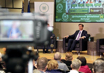 A man in a black chair sits on a stage as a TV camera is pointed at him in the foreground.