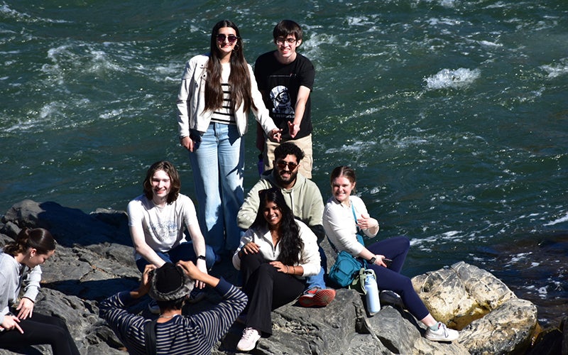 A group of students sit on a rock next to a river.