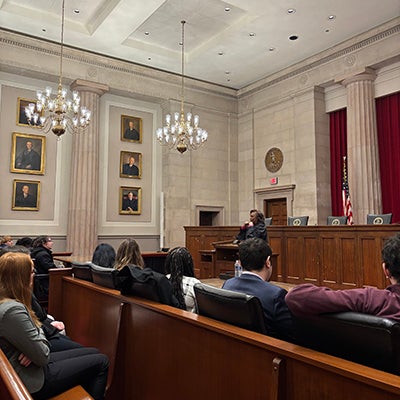 A group of students seated in rows listen to a woman wearing a black robe standing at a podium.