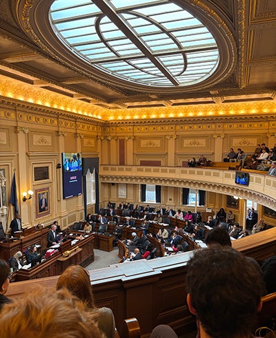 From a balcony, a group of students watch legislators working below in an ornately decorated room.