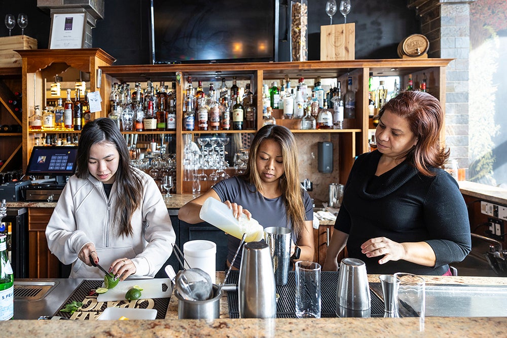 Behind a bartop, a student cuts a lime, another pours a drink, as the supervisor observes