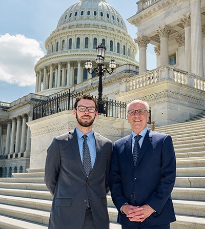 Two men in dark suits and ties stand in front of the Capitol Dome in Washington, D.C.