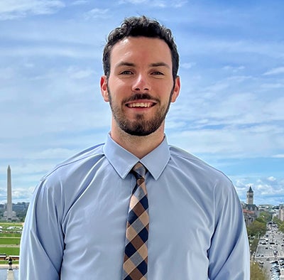 A man in a blue shirt and striped tie stands on a balcony overlooking the Washington Mall with the Washington Monument in the background.