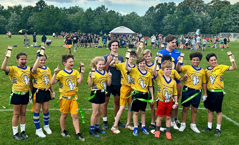 A group of kids wearing yellow flag football shirts stand in front of a man wearing a blue shirt and wave.