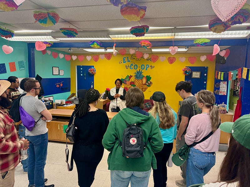 A woman in a black long-sleeved shirt and white and black vest stands in front of a bright yellow wall with the words ‘El Co-op’ and a bowl of fruit painted on it and talks to a group of students.