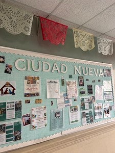 A light blue bulletin board with the words ‘Ciudad Nueva’ hangs below colored lace flags.