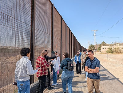 A group of people stand next to a high see-through wall while a few take photos with their phones.