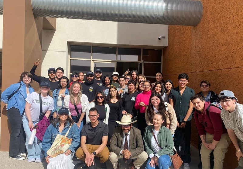 A large group of men and women pose for the camera in front of a university building.
