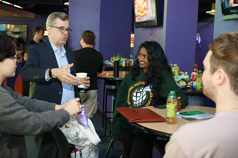 Three students sit listening to a man wearing a blue blazer and blue shirt and holding a cup of coffee.
