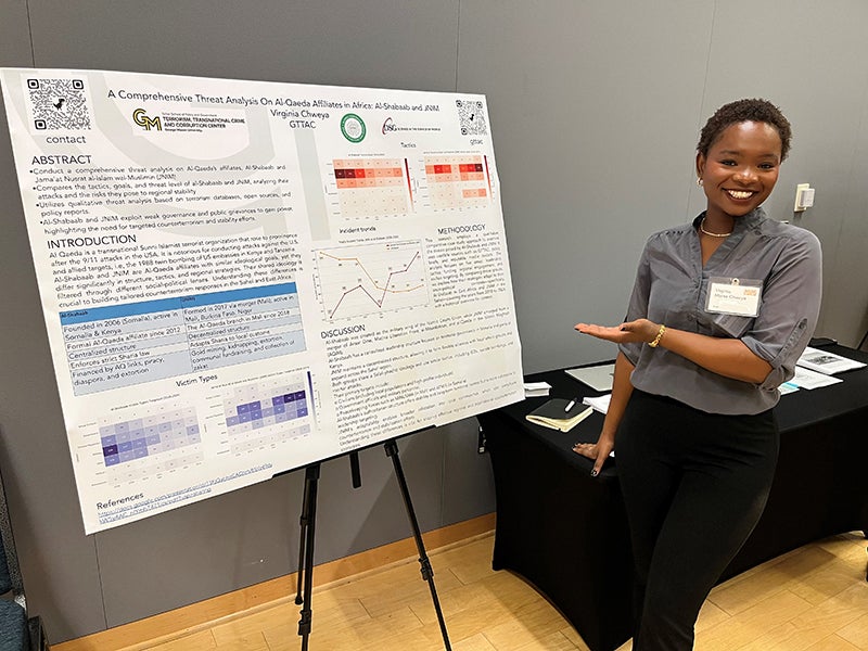 A woman in a gray top gestures to a poster on an easel.