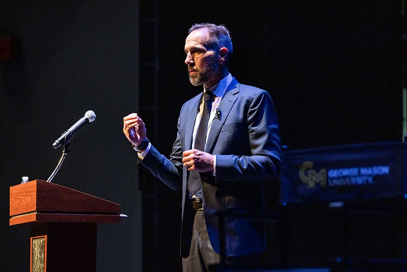 A man in a blue suit jacket stands in front of a microphone.