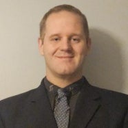A man in a black blazer, black collared shirt, and gray tie stands and smiles.