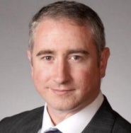 A man in a black blazer, white collared shirt, and tie stands and smiles.