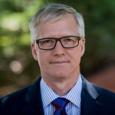 A man wearing a blazer, blue and white striped collared shirt, blue tie, and glasses stands outside and smiles.