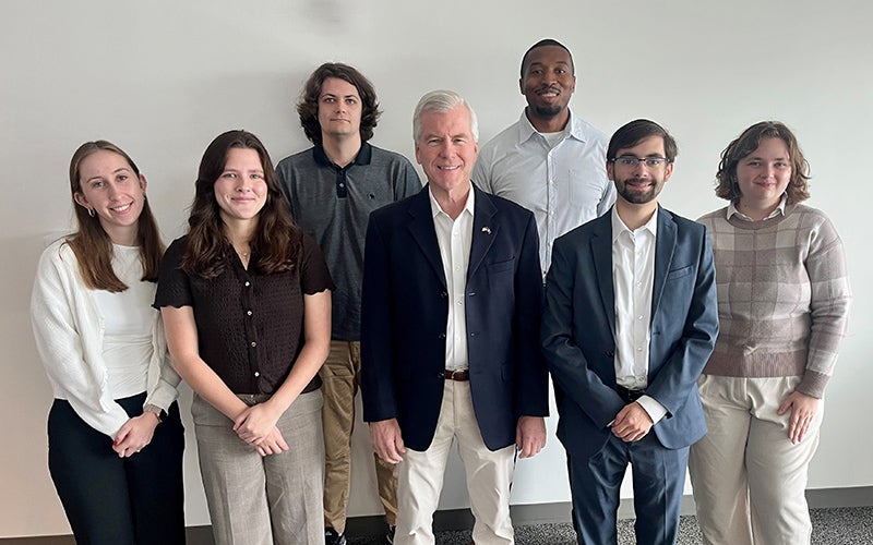 A group of people stand against a white wall and smile at the camera.