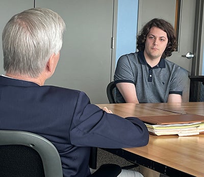 A man with brown hair sits at a table facing a man with white hair.