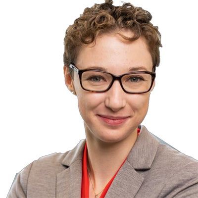 A woman with a brown blazer, red shirt, glasses, and short brown hair stands and smiles
