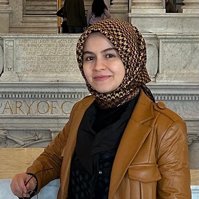 A woman in a brown jacket, black shirt, and head scraf stands outside and smiles