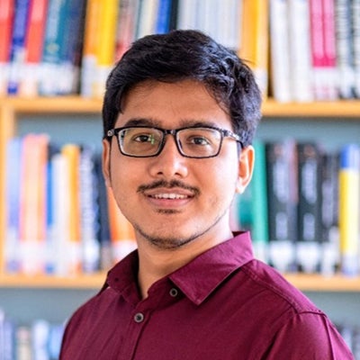 A man in a red collared shirt stands in front of a book case and smiles