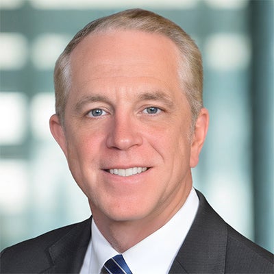 A man in a gray suit, white collared shirt, and tie stands and smiles