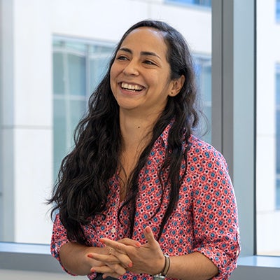 A woman with long back hair and a pink shirt stands inside and smiles