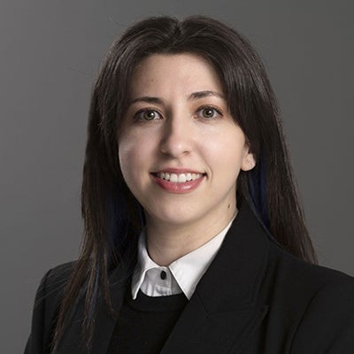 A woman in a black jacket, white collared shirt and long black hair stands inside and smiles