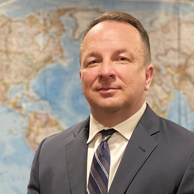 A man in a gray blazer, cream colored shirt, and tie stands in front of a map and smiles