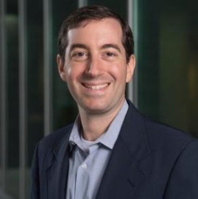 A man in a navy-blue blazer and gray collared shirt stands outside and smiles.